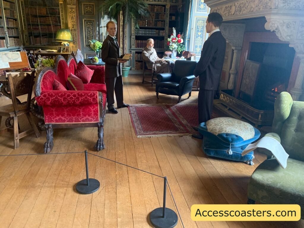 Library State Room with tall bookcases, polished wooden floor, red upholstered sofas, a grand piano, and wax figures dressed in Victorian clothing, with rope barriers marking the viewing area