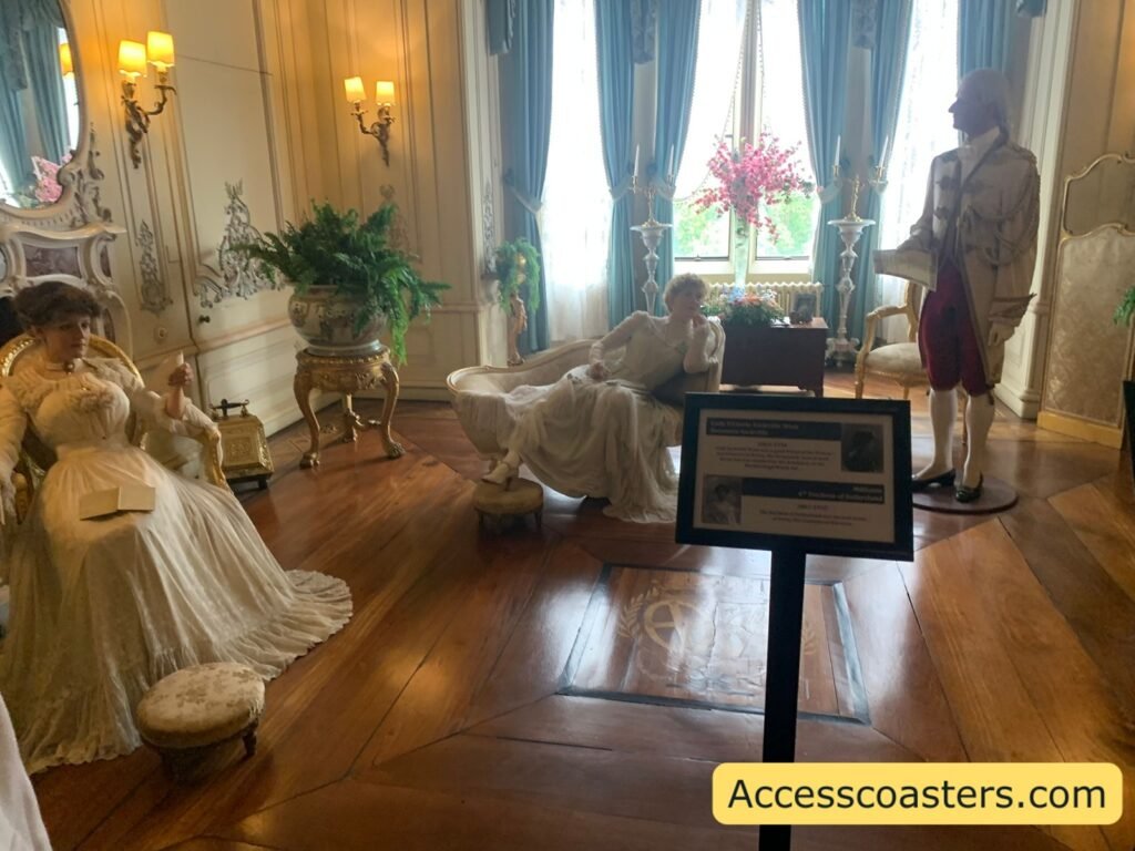 Elegant drawing room State Room with pale walls, chandeliers, tall blue curtains, polished wooden floor, and wax figures in white Victorian clothing reclining on sofas and chairs behind rope barriers, illustrating one of the accessible display scenes.