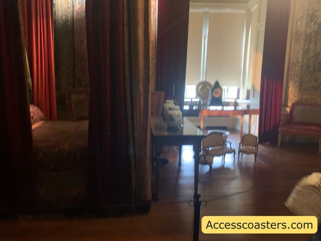 Historic bedroom scene with a large four‑poster bed surrounded by heavy curtains, wooden floor, chairs and a small table near a bright window, all set behind a rope barrier to prevent visitors entering the set.
