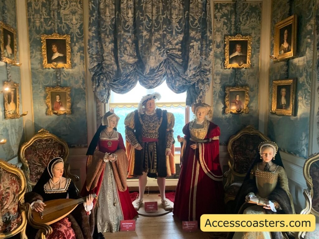 State Room recreated as a Tudor sitting room with blue patterned walls, heavy draped curtains, ornate gold chairs and sofa, and wax figures dressed in rich Medieval and Tudor‑style clothing posed in conversation.
