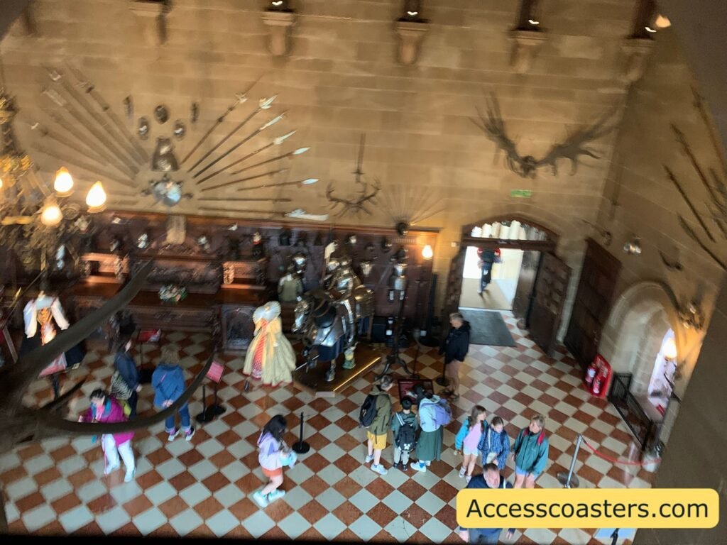 Wide view of the Great Hall with a chequered red‑and‑cream floor, long wooden tables, displays of arms and armour on the walls, antlers above, and visitors