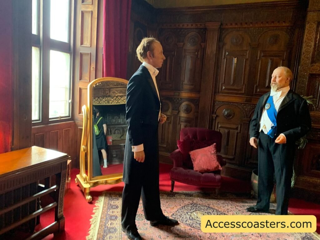 Royal Weekend Party waxwork scene at Warwick Castle showing two formally dressed male figures in a richly decorated state room, one in a black tailcoat and the other wearing ceremonial dress with a blue sash, standing on a patterned rug in front of dark wood panelling and tall windows.