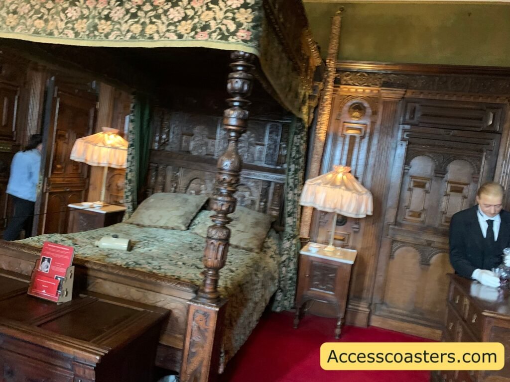 : Royal Weekend Party bedroom scene at Warwick Castle featuring a huge carved four-poster bed with an ornate canopy, patterned green bedding and fringed bedside lamps, set against richly panelled wooden walls.