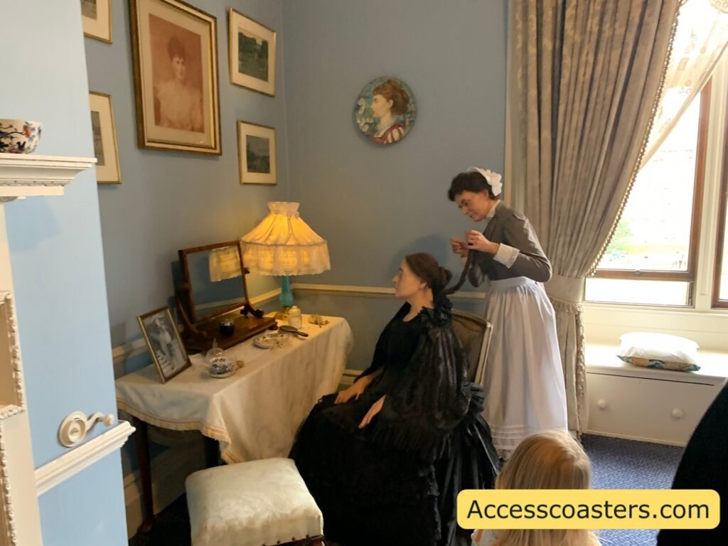 Royal Weekend Party waxwork scene of a Victorian-style bedroom or dressing room at Warwick Castle, with a seated woman in a dark dress at a dressing table while a lady’s maid in a long grey dress stands behind her, surrounded by framed pictures and soft lamplight.