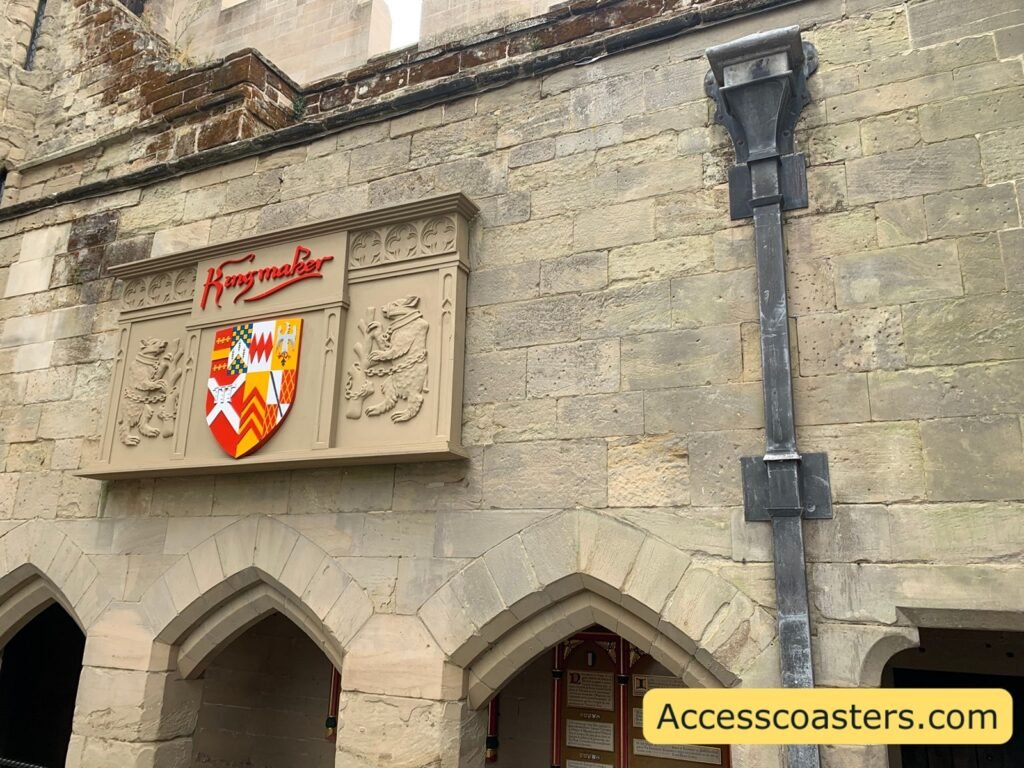  Exterior stone façade of The Kingmaker at Warwick Castle, with arched doorways and colourful heraldic shields mounted above, showing the medieval theming of the attraction.
