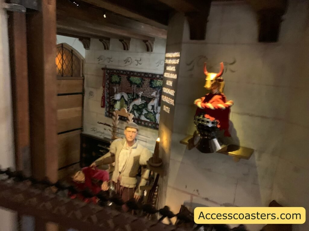 Overhead view into a busy interior scene from The Kingmaker, with a costumed wax figure speaking below and another figure in bright red and gold armour on a raised platform, plus projected text on the wall