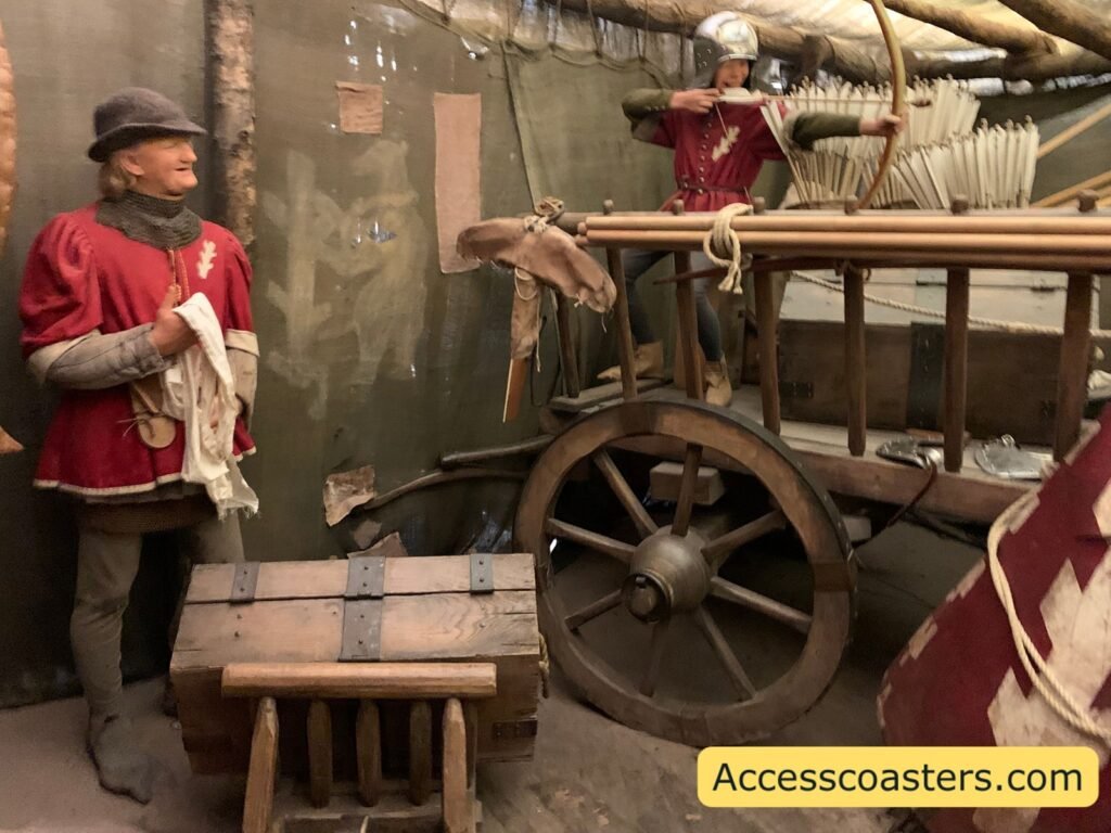  Armoury and supply area scene in The Kingmaker, with a wax figure in period clothing standing beside a wooden cart loaded with equipment, barrels and bundles of arrows ready for the War of the Roses.