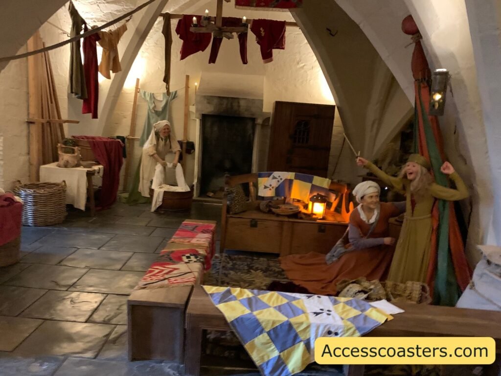 Medieval bedroom or campaign tent scene in The Kingmaker, with a stone floor, wooden chests, a colourful chequered bedspread and costumed wax figures preparing clothes and equipment for war.
