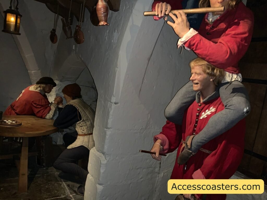  Dimly lit medieval kitchen or workroom scene inside The Kingmaker, with realistic wax figures in red livery talking and working around a stone archway and wooden table, showing the immersive theming.