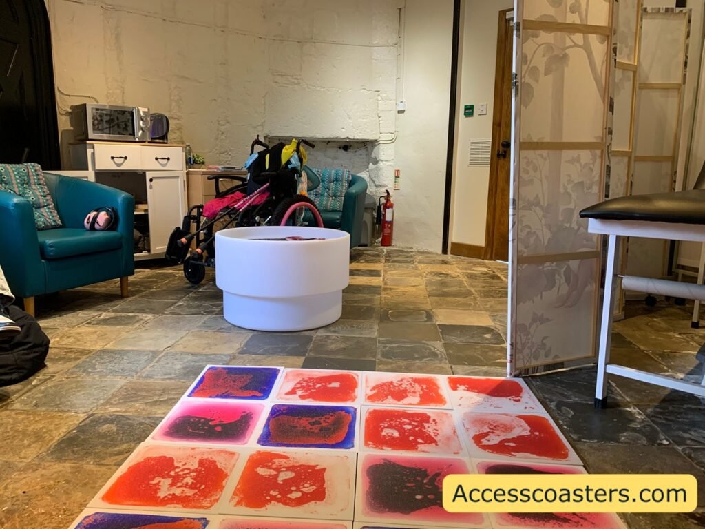 Wider view of the Welfare and Sensory Room showing the beanbag corner, a teal armchair, a white tub or ball pit, spare clothes and bags, and colourful liquid-filled floor tiles on the stone floor