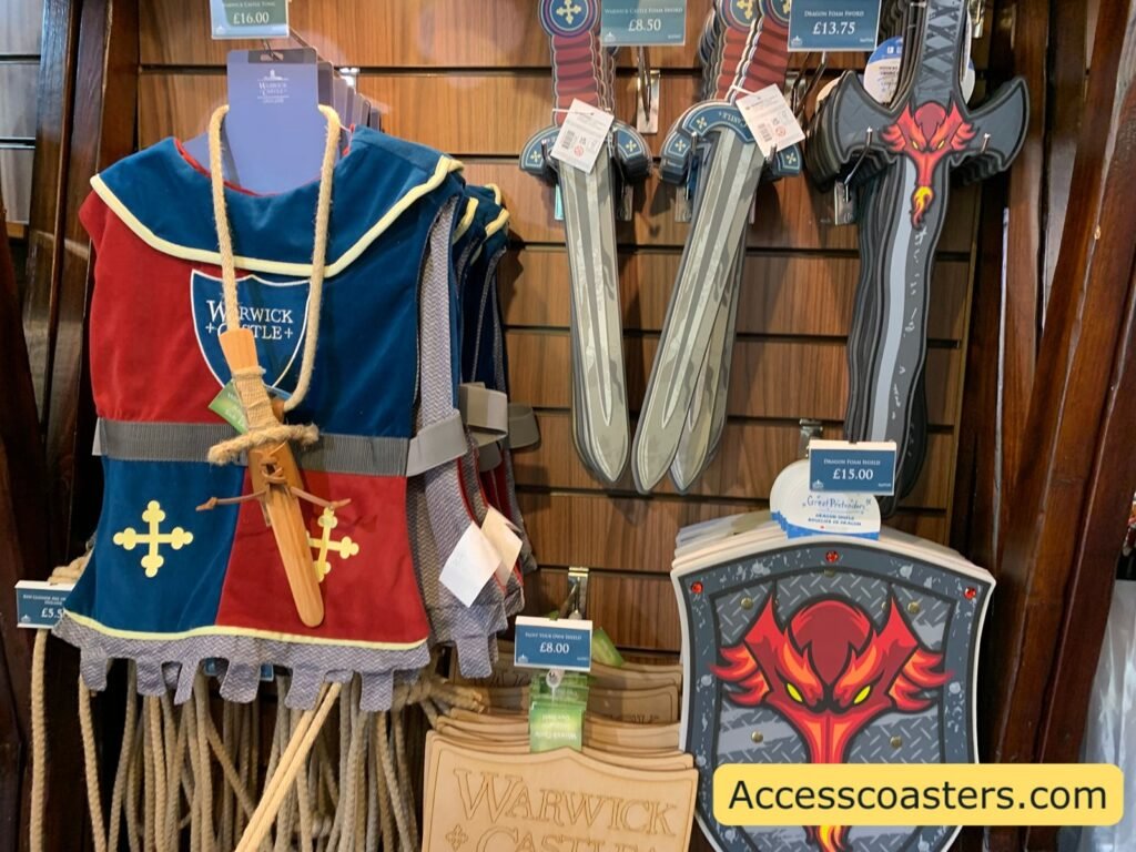 Display of Warwick Castle branded children’s knight costumes in a gift shop, including a blue tunic with a gold cross, toy swords, shields and belts hanging on a wooden wall.
