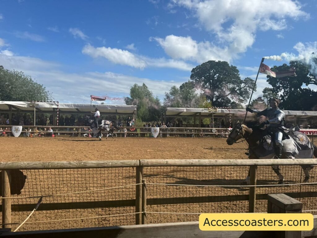 Wide view across the Wars of the Roses Live jousting arena, with a knight on horseback charging along the sand track while holding a lance and flag, in front of covered spectator stands.
