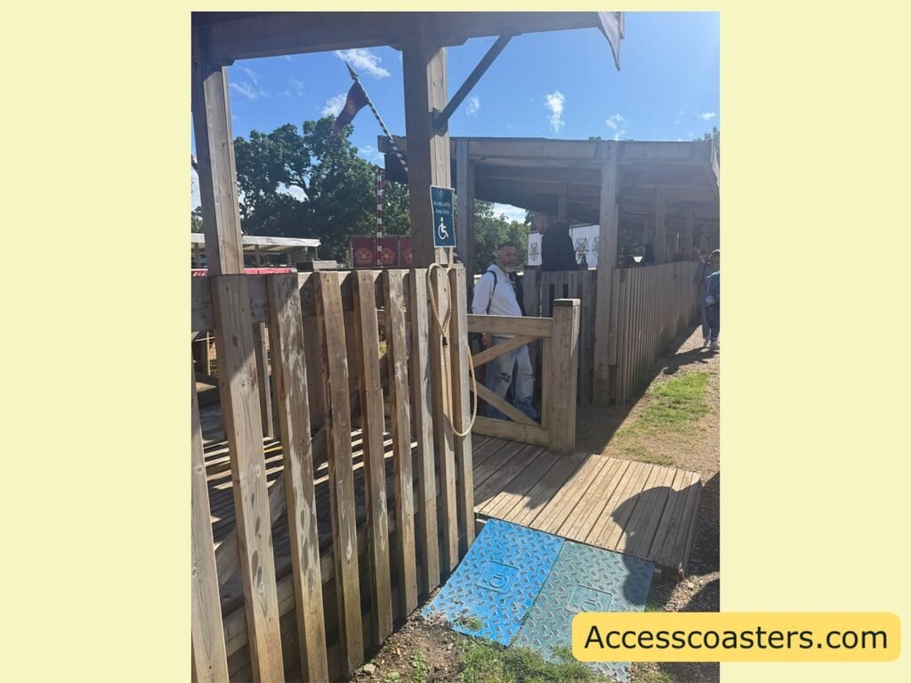 Accessible viewing area entrance for the Wars of the Roses Live arena, showing wooden fencing, a ramp with a blue non-slip mat, and a covered standing platform where guests can watch the show.
