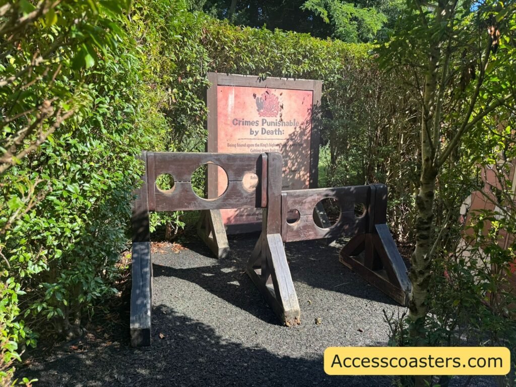  A wooden stock-style photo point in the maze, with three round holes at head height in a dark wooden frame, set between hedge walls and a sign behind it.