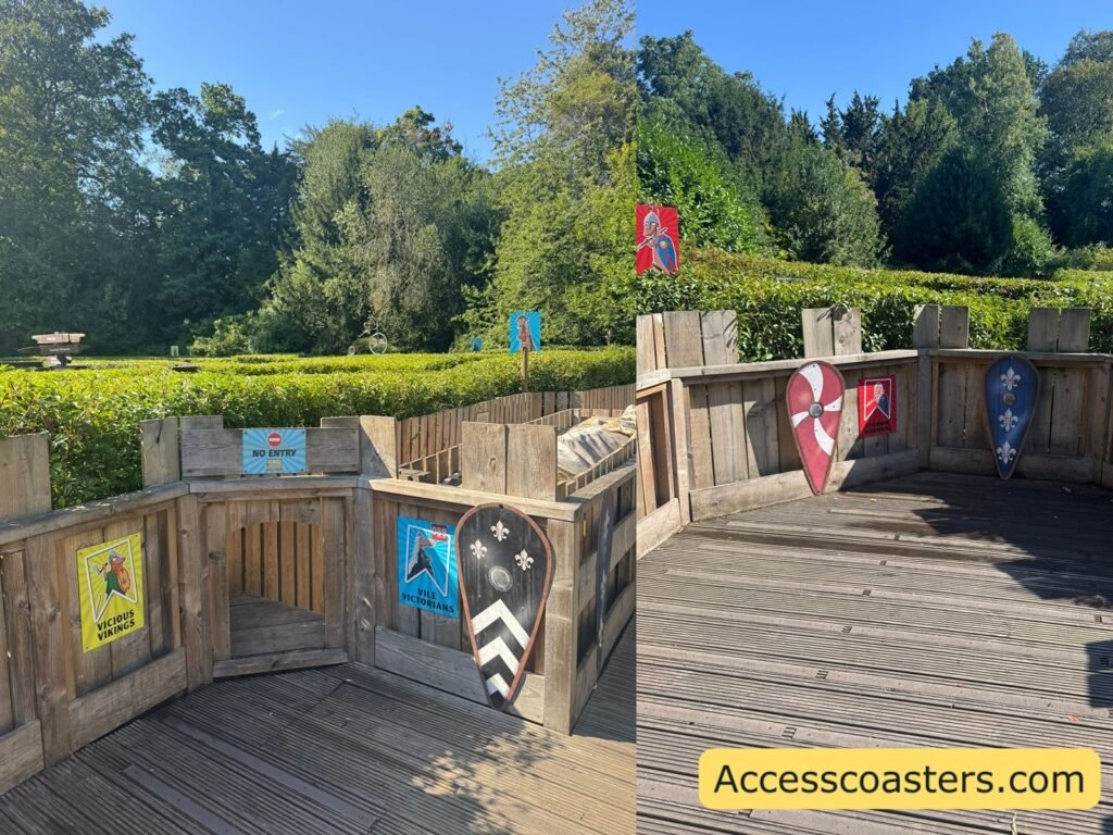  A raised wooden viewing platform with railings and several colorful “Horrible Histories” character boards fixed around the sides, looking out over more hedges and trees.