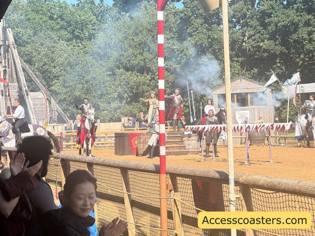Busy scene in the Wars of the Roses Live arena during the battle finale, with multiple riders, actors, striped poles, smoke and special effects, and a cheering crowd watching from behind the barrier.