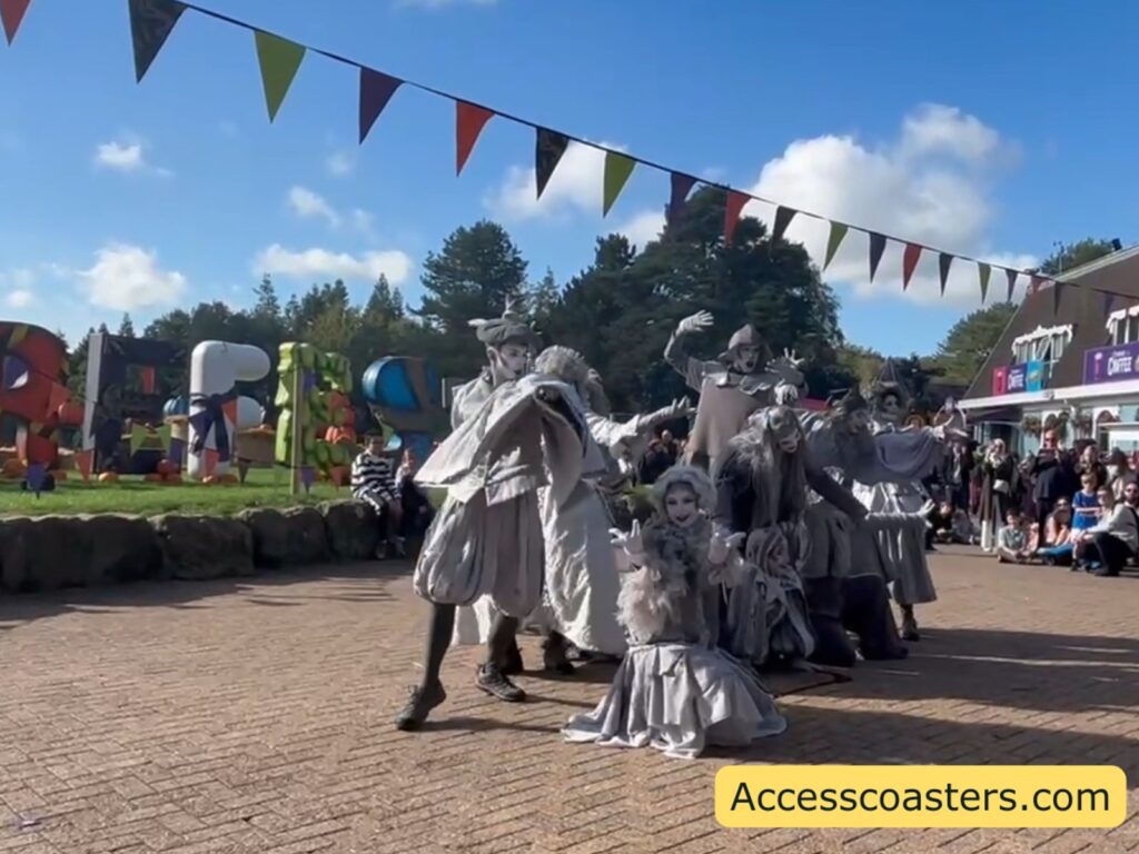 A group of performers dressed as ghostly, grey Victorian‑style “statue” characters dance in formation on the main path, framed by colourful bunting overhead. Guests watch from the sides and in the background