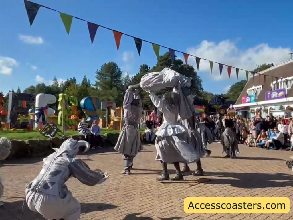 Close‑up moment in the Alton Ancestors flash mob on Towers Street. Several grey, statue‑like dancers strike dramatic poses mid‑routine, including one performer crouched low at the front while others move behind them. The action happens on the open paving with passers‑by  visible 