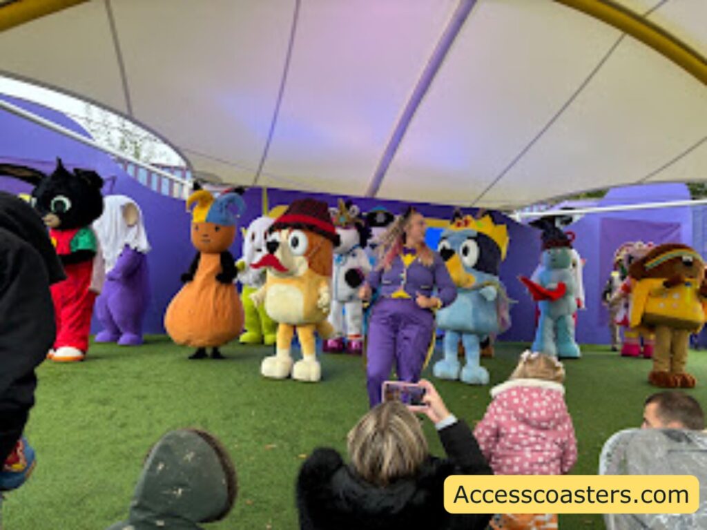 Closer view of the Monster Ball stage under the canopy, showing a full line‑up of costumed CBeebies friends in their Halloween outfits standing on the grass in front of a purple backdrop while children and adults sit close to the front, some holding up phones to record the spooky dance.