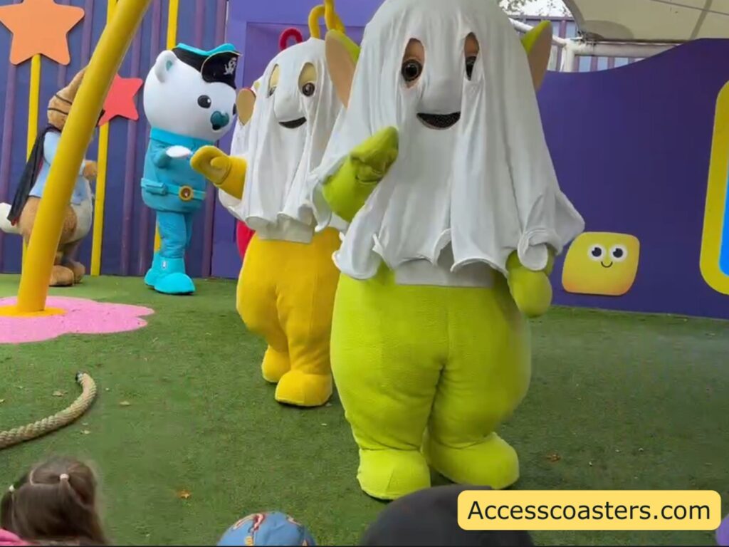 Close‑up of the Monster Ball parade on the grass, with two Teletubbies in bright yellow and green ghost costumes walking toward the audience and more colourful CBeebies characters in Halloween outfits standing behind them