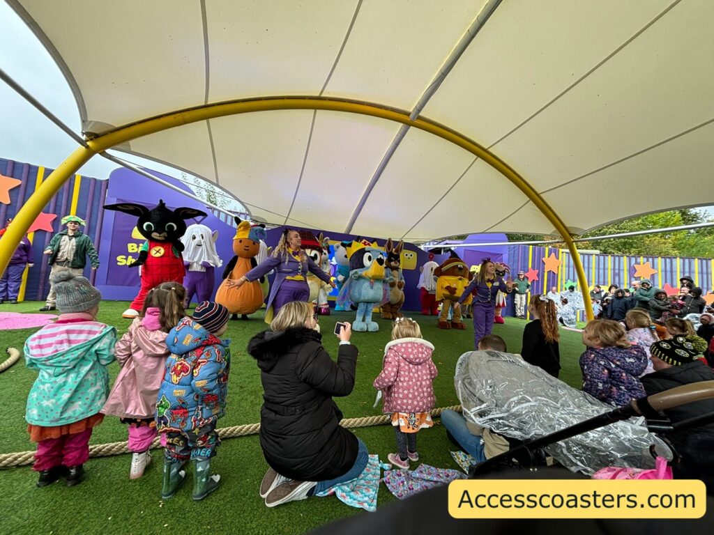 Wide shot of the CBeebies Land Monster Ball show in the Big Fun Showtime area at Alton Towers, with rows of children and adults sitting on artificial grass under a large white canopy, all facing a stage where lots of brightly coloured CBeebies costume characters in Halloween outfits dance and wave 