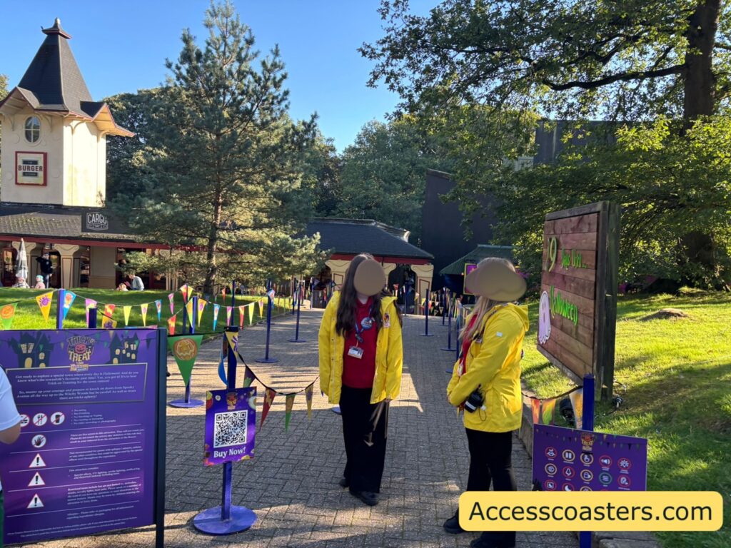 Entrance to Trick O’ Treat Town at Alton Towers in bright afternoon light, with two staff members in yellow jackets standing beside a purple information board and queue barrier at the start of the path, with themed buildings and trees in the background.