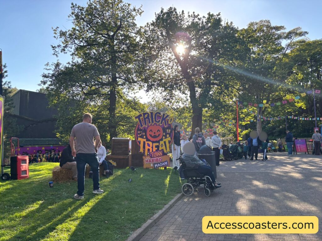 Pathway in Fountain Square at Alton Towers leading towards the colourful circular Trick O’ Treat Town entrance sign, surrounded by grass  with lots of prams parked outside and people gathered 